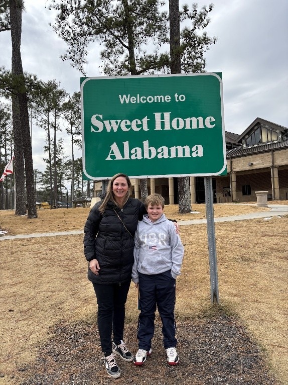 Woman and boy pose by a "Welcome to Sweet Home Alabama" sign in a wooded area, capturing a joyful moment during their visit.
