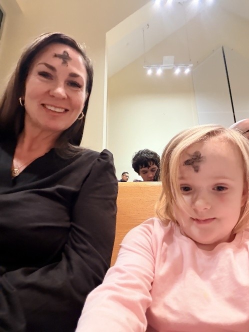 A woman and a young girl sit together, both with ash crosses on their foreheads, indicating Ash Wednesday. They smile in a church setting.