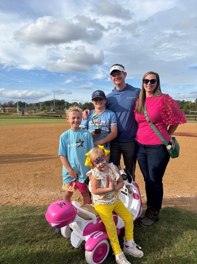A family celebrates a youth sports achievement on a baseball field. The boy in the middle holds a trophy, while siblings and parents smile, with one child on a toy motorcycle.