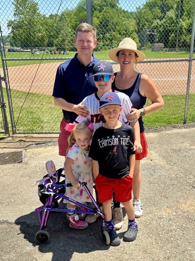 Family of five poses together at a baseball field, with children in sports attire and a young girl in a stroller. Bright, sunny day.
