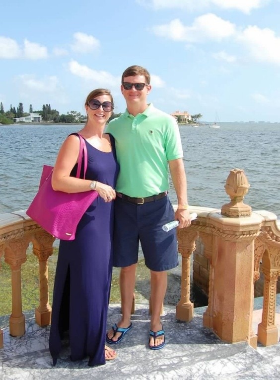 A couple poses together by the waterfront, smiling under a sunny sky. The woman wears a long navy dress and carries a pink bag, while the man is in a green polo and shorts.