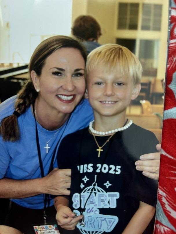 A smiling woman with long hair poses with a boy wearing a black shirt and pearl necklace, both appearing joyful in a classroom setting.