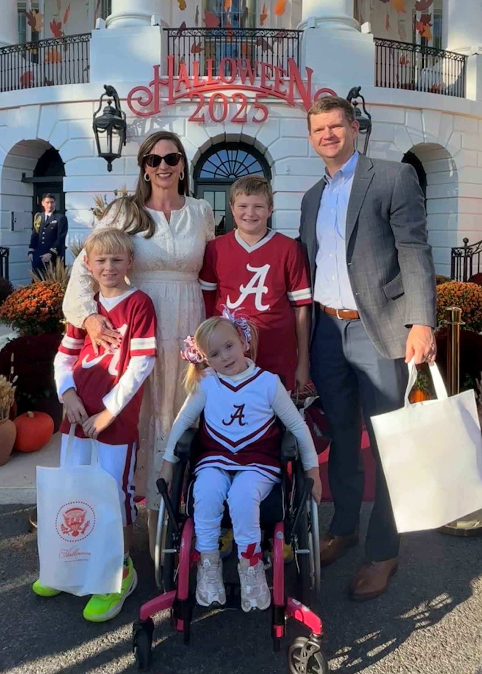 A family poses together outside, celebrating Halloween 2025. Children wear Alabama-themed outfits, and the scene features festive decorations.