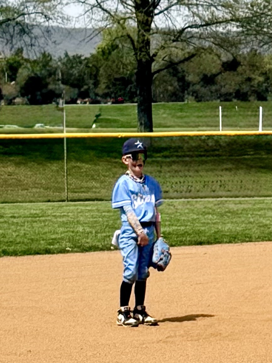A young baseball player in a light blue uniform stands on a sunny field, smiling with excitement. The background features a tree and outfield fence.