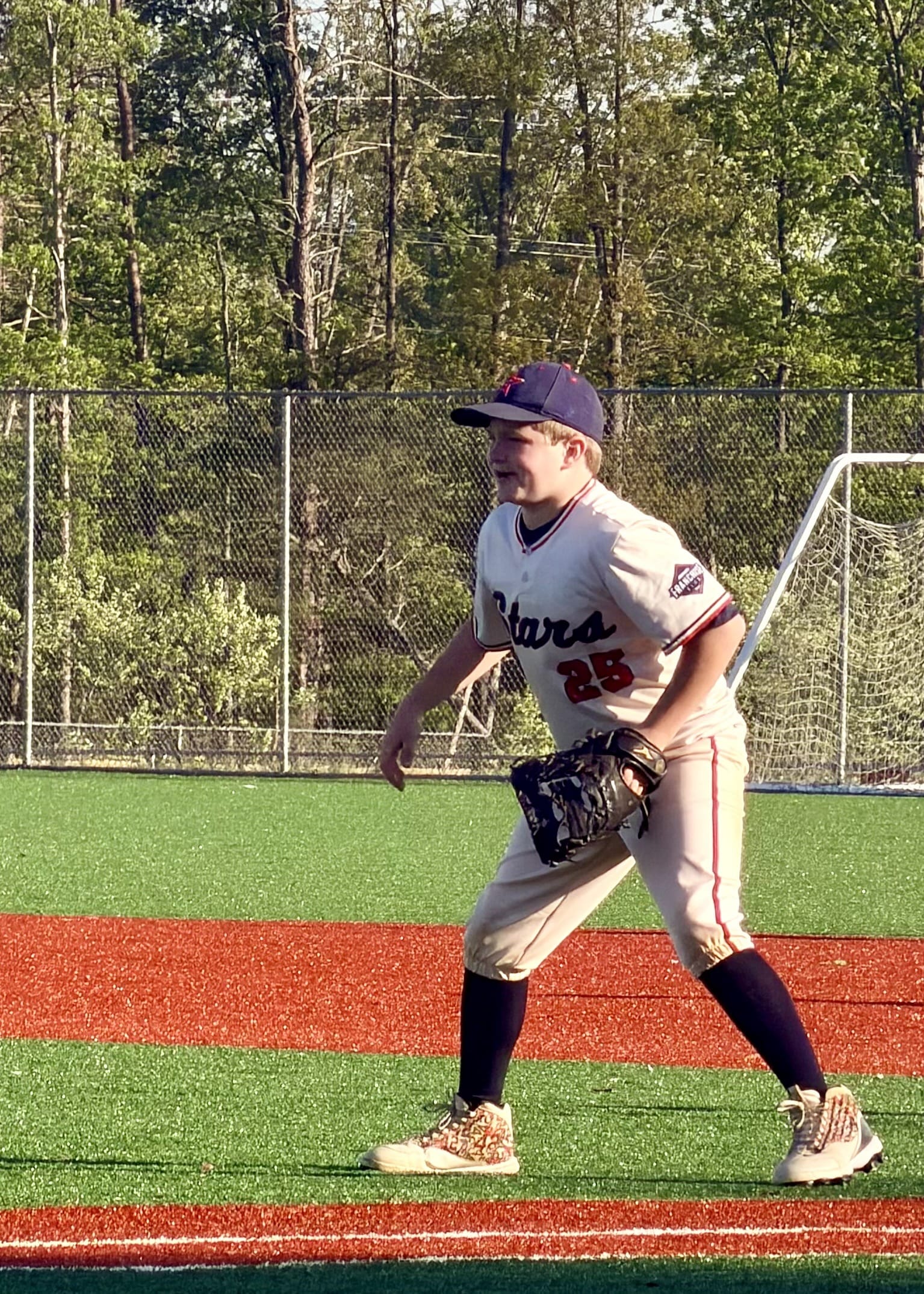 A young baseball player in a cream uniform and navy cap stands ready on a vibrant green and red field, with a focused expression and trees in the background.